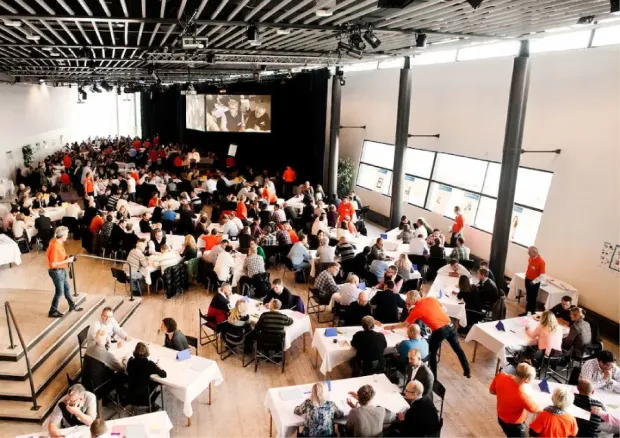 A large group of people are seated at tables in a brightly lit conference hall, with some individuals standing and interacting playing a type of business simulation